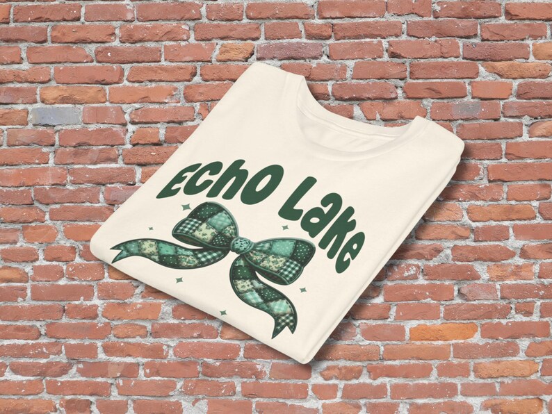 May include: Cream-colored t-shirt featuring the text "Echo Lake" in dark green, with a plaid bow graphic in shades of green and teal. The shirt is folded and displayed against a brick wall.