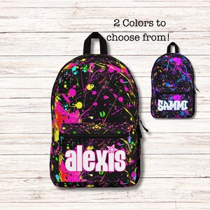May include: Black backpack with a colorful paint splatter design in pink, yellow, and blue. The backpack has the name "alexis" in white block letters. A smaller backpack with the name "SAMMI" is also shown.