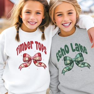 May include: Two young girls wearing sweatshirts. One sweatshirt is white with the text "Timber Tops" and a red and pink bow graphic. The other is gray with "Echo Lake" and a green bow graphic.