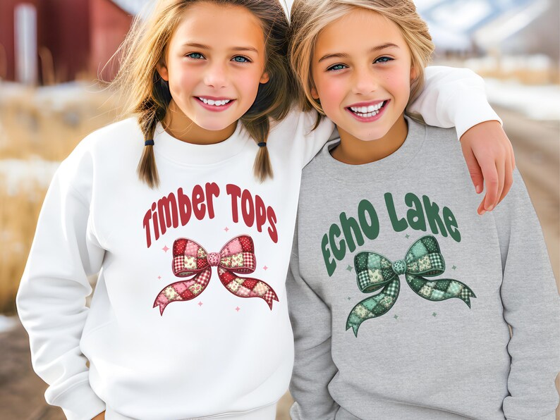 May include: Two young girls wearing sweatshirts. One sweatshirt is white with the text "Timber Tops" and a red and pink bow graphic. The other is gray with "Echo Lake" and a green bow graphic.