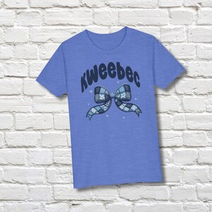 May include: A blue t-shirt with the word "KweeBec" in a curved font above a bow graphic. The bow is a patchwork design in shades of blue and white. The shirt is displayed against a white brick wall.