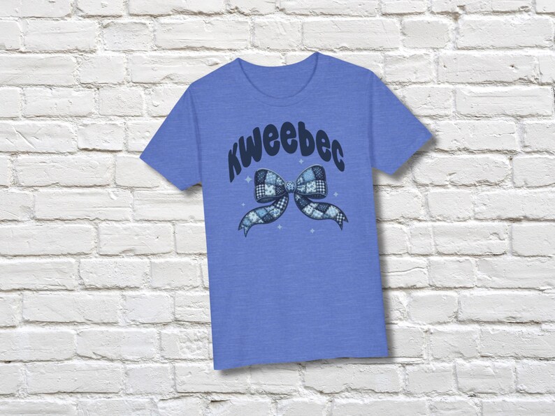 May include: A blue t-shirt with the word "KweeBec" in a curved font above a bow graphic. The bow is a patchwork design in shades of blue and white. The shirt is displayed against a white brick wall.