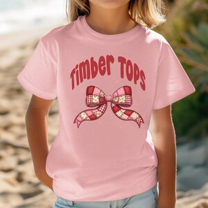 May include: A light pink t-shirt with the words "Timber Tops" in red script above a patchwork bow design. The bow features red, pink, and white patterned fabric. The shirt is short-sleeved.