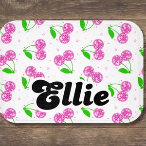 May include: A white rectangular bath mat featuring a pattern of pink disco ball cherries with green stems and leaves, and small stars. The name "Ellie" is printed in black, stylized font across the mat.