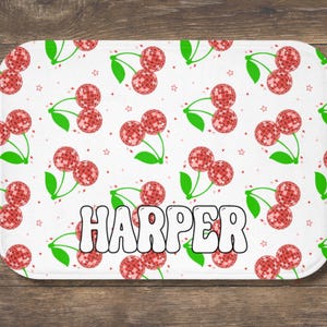 May include: A white rectangular bath mat with a repeating pattern of red disco ball cherries and green leaves. The name "HARPER" is printed in large, white, bubble letters across the bottom of the mat.
