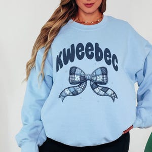 May include: Light blue sweatshirt with the word "kweeebec" in dark blue script above a patchwork bow design. The bow features various blue patterns. The sweatshirt is a solid light blue color.
