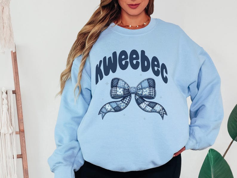 May include: Light blue sweatshirt with the word "kweeebec" in dark blue script above a patchwork bow design. The bow features various blue patterns. The sweatshirt is a solid light blue color.