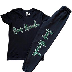 Puede incluir: Conjunto de camiseta y pantal&oacute;n de ch&aacute;ndal negro y verde con el texto "Camp Monasha" en una fuente verde de escritura.