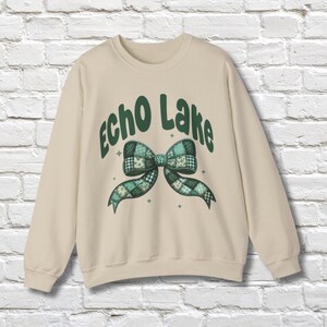 May include: A cream-colored sweatshirt with the words "Echo Lake" in green script above a green and blue plaid bow design. The bow has a patchwork pattern and is centered on the front of the shirt. The sweatshirt has long sleeves.