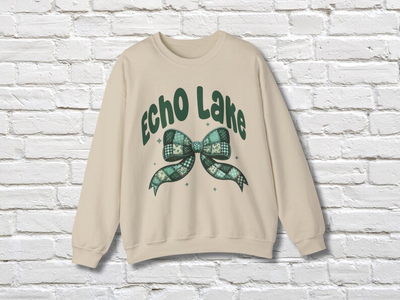 May include: A cream-colored sweatshirt with the words "Echo Lake" in green script above a green and blue plaid bow design. The bow has a patchwork pattern and is centered on the front of the shirt. The sweatshirt has long sleeves.