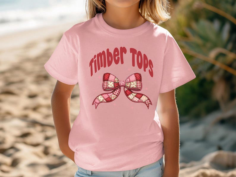 May include: A light pink t-shirt with the words "Timber Tops" in red script above a patchwork bow design. The bow features red, pink, and white patterned fabric. The shirt is short-sleeved.