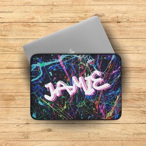May include: A black laptop sleeve with a colorful paint splatter design. The name "JAMIE" is written in white with a pink outline.