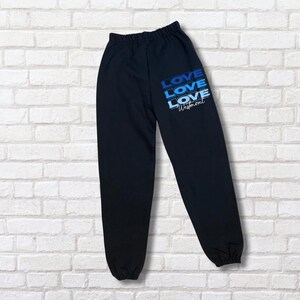 May include: Black sweatpants with an elastic waistband and cuffs. The text "LOVE LOVE LOVE Westmont" is printed in blue gradient lettering on the left leg. The pants are made of a soft material.