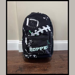 May include: Black backpack with a football design in white and green accents. The backpack features a front pocket with the name "ZOPPE" in green. The design includes a football and splatter effects.
