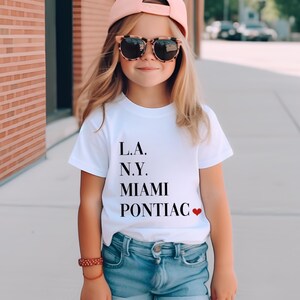 May include: White t-shirt with black text that reads "L.A. N.Y. MIAMI PONTIAC" with a red heart after the last word. A young person is wearing the shirt with blue denim shorts, a pink baseball cap, and sunglasses.