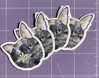 May include: Three black, white and orange cat stickers with green eyes. The stickers are on a purple gridded surface.
