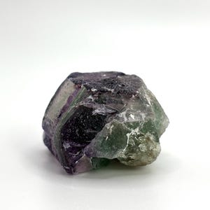 May include: A raw, natural fluorite crystal with a mix of colors. The stone features shades of purple, green, and clear sections, with a rough, textured surface. The crystal is set against a white background.
