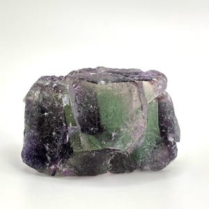 May include: A raw fluorite crystal with a mix of purple, green, and clear sections. The natural mineral has a rough, textured surface and irregular shape. The crystal is set against a white background.