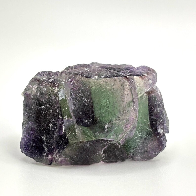 May include: A raw fluorite crystal with a mix of purple, green, and clear sections. The natural mineral has a rough, textured surface and irregular shape. The crystal is set against a white background.