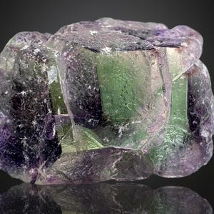 May include: A close-up of a fluorite mineral specimen. The crystal formation displays a mix of colors, including green, purple, and clear sections. The surface has a reflective quality, with a dark background.