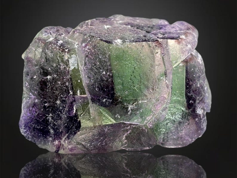 May include: A close-up of a fluorite mineral specimen. The crystal formation displays a mix of colors, including green, purple, and clear sections. The surface has a reflective quality, with a dark background.