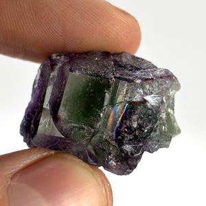 May include: A close-up of a raw fluorite crystal. The crystal displays a mix of colors, including purple, green, and hints of rainbow iridescence. The rough texture and natural form of the mineral are visible.