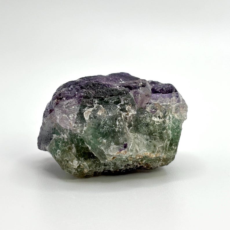 May include: A rough, uncut fluorite crystal with a mix of purple, green, and clear sections. The natural stone has a textured surface and irregular shape, set against a white background.