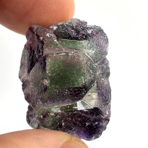 May include: A raw, uncut fluorite crystal with a mix of purple, green, and clear sections. The stone has a rough, natural texture and is held between two fingers against a white background. The crystal's facets catch the light, showcasing its color variations.