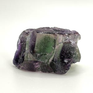 May include: A raw fluorite crystal with a mix of purple, green, and clear sections. The natural mineral has a rough, textured surface and a crystalline structure. The stone is set against a white background.