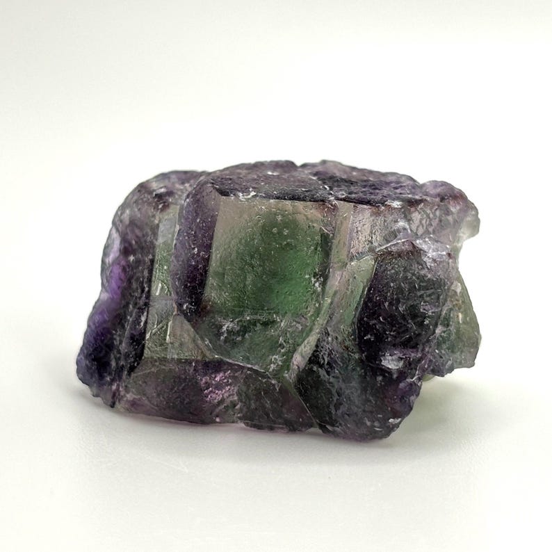 May include: A raw fluorite crystal with a mix of purple, green, and clear sections. The natural mineral has a rough, textured surface and a crystalline structure. The stone is set against a white background.