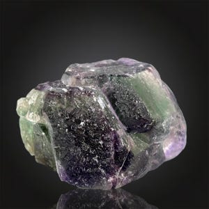 May include: A close-up of a raw fluorite crystal. The crystal displays a mix of colors, including deep purple, green, and clear sections. The surface has a rough, natural texture, with visible crystalline structures. The lighting highlights the mineral's natural beauty.