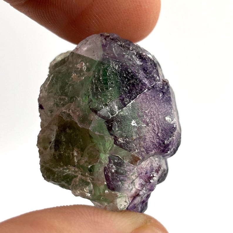 May include: A raw, natural fluorite crystal with a mix of colors. The crystal features shades of purple, green, and clear sections. The rough texture and irregular shape of the stone are visible. The crystal is held between two fingers against a white background.