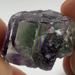 May include: A close-up of a fluorite crystal with a mix of colors. The crystal has a dark gray exterior with visible facets and a translucent interior. A rainbow effect is visible within the crystal, with shades of green, purple, and blue.