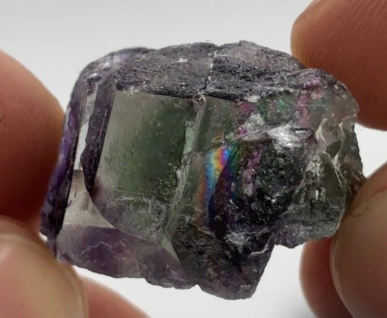 May include: A close-up of a fluorite crystal with a mix of colors. The crystal has a dark gray exterior with visible facets and a translucent interior. A rainbow effect is visible within the crystal, with shades of green, purple, and blue.