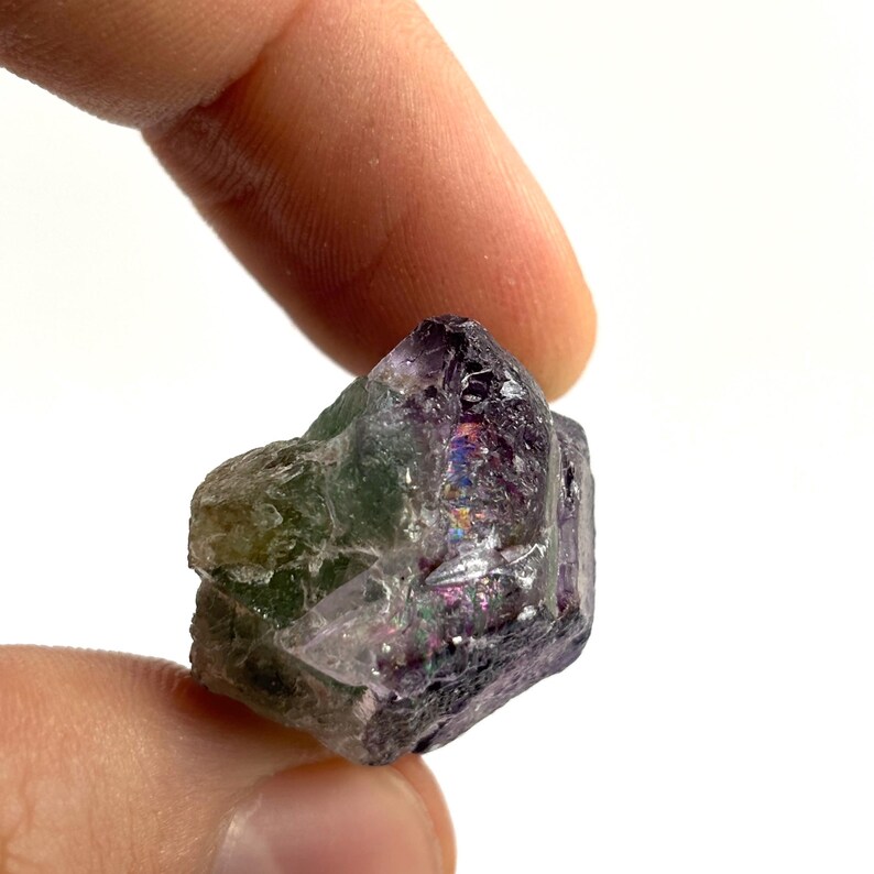 May include: A small, raw fluorite crystal held between fingers. The crystal displays a mix of colors, including purple, green, and hints of iridescent hues. The rough texture and natural form are visible against a white background.