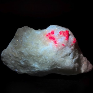 Puede incluir: Primer plano de una roca blanca con depósitos minerales de color rojo brillante. La roca tiene una superficie rugosa y texturizada y se encuentra sobre un fondo negro. Los minerales rojos están agrupados en la parte superior de la roca.