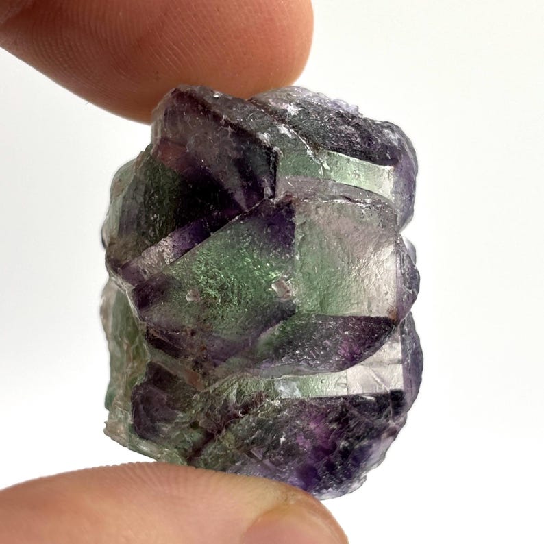 May include: A close-up of a raw fluorite crystal. The crystal displays a mix of colors, including purple, green, and clear sections. The rough texture and natural form of the mineral are visible. The crystal is held between two fingers.