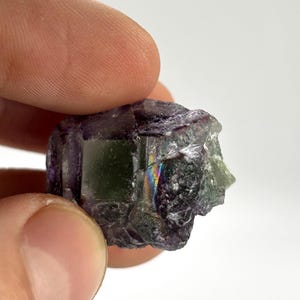 May include: A raw, natural fluorite crystal with a mix of purple, green, and clear sections. The crystal has a rough, textured surface and displays a subtle rainbow iridescence in certain areas. The stone is held between fingers.