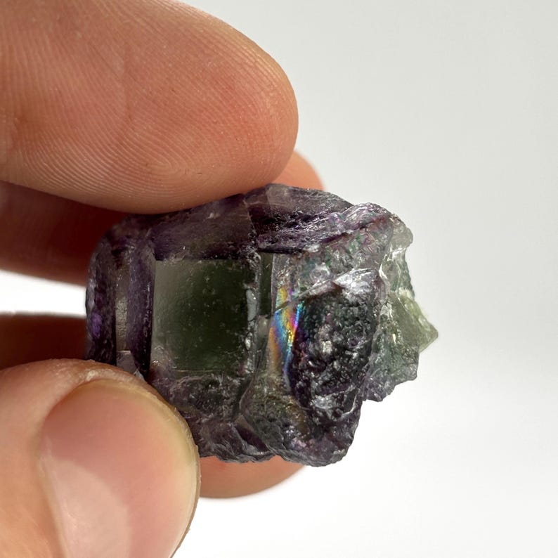 May include: A raw, natural fluorite crystal with a mix of purple, green, and clear sections. The crystal has a rough, textured surface and displays a subtle rainbow iridescence in certain areas. The stone is held between fingers.