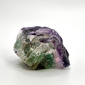 May include: A raw fluorite crystal with a mix of purple, green, and clear sections. The natural stone has a rough, uneven surface and is set against a white background. The crystal's colors are vibrant and well-defined.