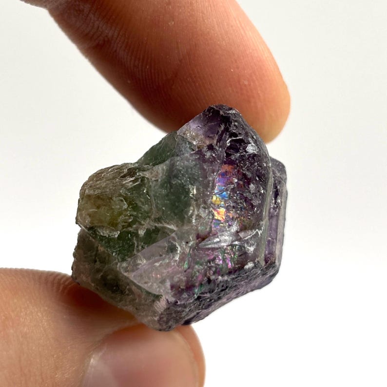 May include: A close-up of a raw, uncut fluorite crystal. The crystal displays a mix of colors, including green, purple, and iridescent patches. The rough texture and natural form of the stone are visible, with the light reflecting off the facets.
