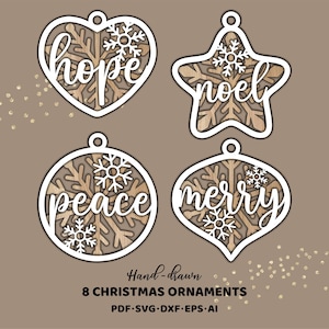 Op de afbeelding: Vier houten kerstversieringen in hart-, ster-, cirkel- en druppelvorm. Elk ornament heeft een witte omtrek en bevat de woorden: "hope", "noel", "peace" en "merry". De ornamenten hebben een sneeuwvlokontwerp en zijn met de hand getekend.