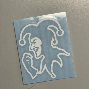 Op de afbeelding: Witte sticker met een narrengezicht op een lichtblauwe achtergrond. De nar heeft een lachend gezicht en draagt een hoed met drie punten. De sticker is rechthoekig.