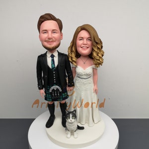 May include: A wedding cake topper featuring a bride and groom in a custom caricature style. The groom is wearing a black suit with a green kilt and the bride is wearing a white dress. A black and white cat is sitting in front of them.