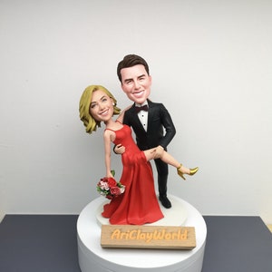 May include: A custom-made figurine of a couple in a wedding pose. The woman is wearing a red dress and the man is wearing a black tuxedo. The figurine is on a white base with a wooden plaque that reads "AriClayWorld".