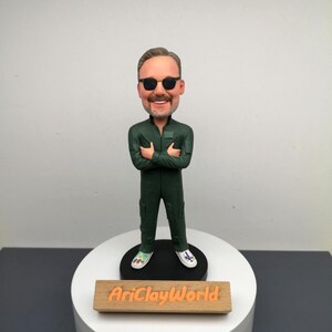 Puede incluir: Una figurita con un mono verde, gafas de sol y zapatos coloridos. La figurita está de pie sobre una base circular blanca con un letrero de madera que dice "AriClayWorld".