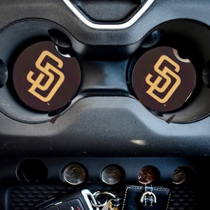 May include: Two black cup holders with brown coasters featuring the San Diego Padres logo in gold. The logo is a stylized "SD" with a baseball inside the "D".