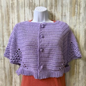 May include: A light purple, short-sleeved, crocheted cardigan with three matching buttons down the front. The cardigan has a scalloped hem and decorative openwork on the sleeves. The garment is displayed on a white mannequin head and torso.