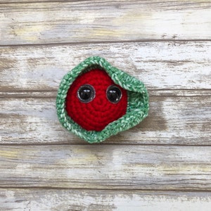 May include: A handmade crochet creature with a red body and large, shiny black eyes. The red body is nestled within a green and white crocheted wrap. The item is placed on a light-colored, wooden surface.