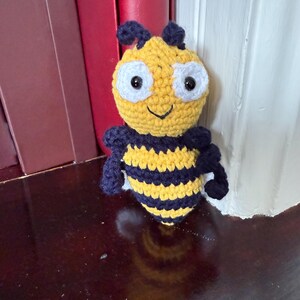 May include: A handmade crochet bee doll with a yellow body, black and yellow striped abdomen, and dark blue wings and antennae. The bee has large white eyes and a smiling face. The doll is sitting on a dark brown surface.